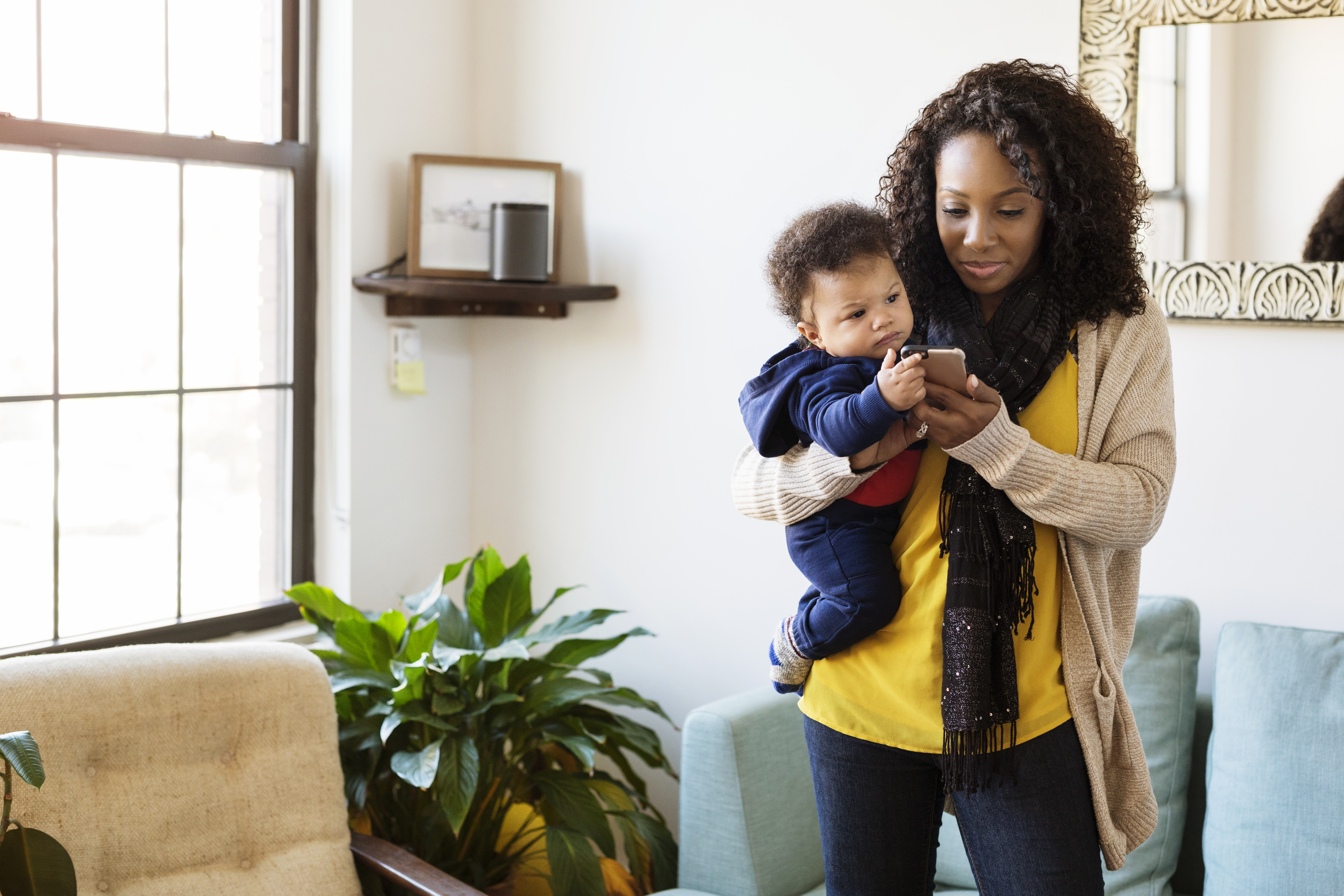 Smiling woman using mobile phone while carrying baby. Mother holding son and texting on cell phone at home.
